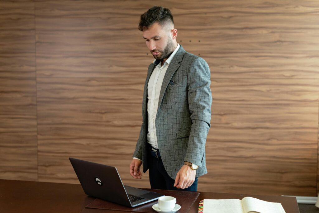 Businessman in gray blazer stands working on laptop at office desk.